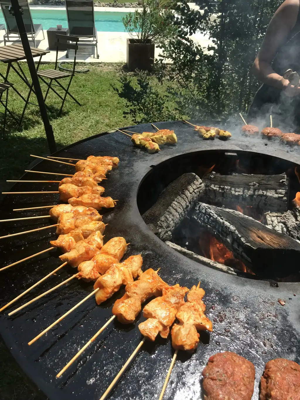 Brochettes de poulet marinées et boulettes de viande cuisent sur une grande plancha barbecue extérieure. Feu de bois et piscine en arrière-plan.
