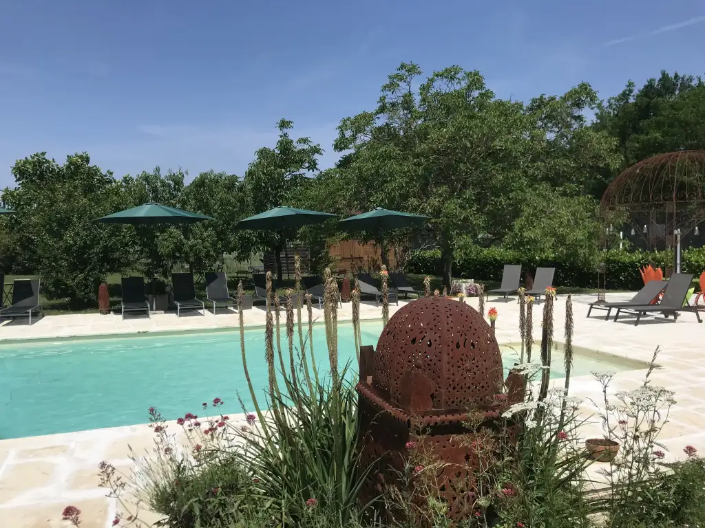 Piscine de luxe avec margelle en pierre, transats et parasols verts. Sculpture décorative en métal rouillé au milieu d'un jardin luxuriant.